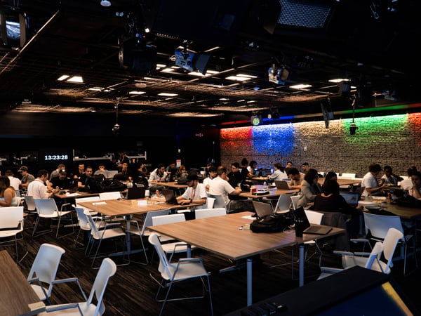 A wide-angle photograph of a large, dimly lit indoor space where numerous people are seated at wooden tables, working on laptops. The room features professional lighting equipment on the ceiling and a back wall illuminated with red, blue, and green lights.