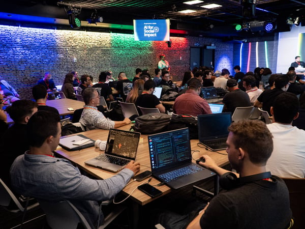 A group of people are seated at long tables with laptops open, attending an 'AI for Social Impact' event hosted by Google Developers.