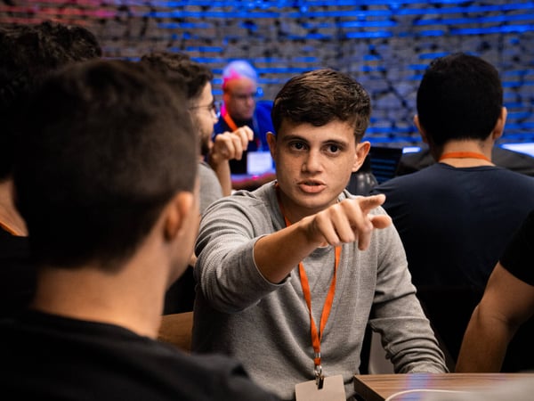 A young man in a grey shirt and orange lanyard points toward someone while engaged in a discussion at an indoor event.