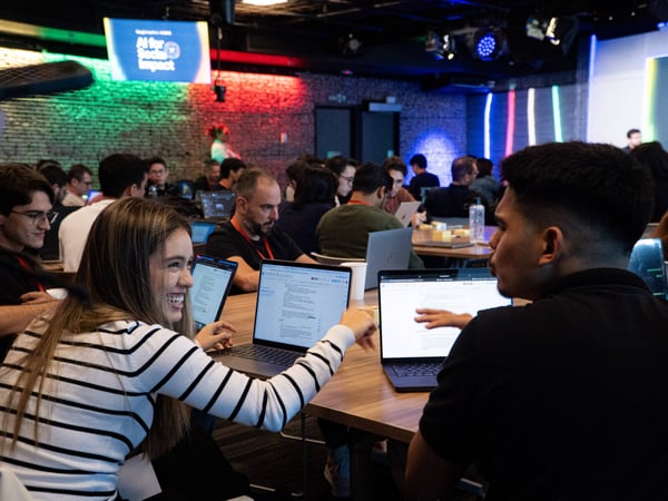 A group of people are gathered in a dimly lit event space, working on laptops and engaging in discussion, with a screen in the background that reads 'AI for Social Impact'.