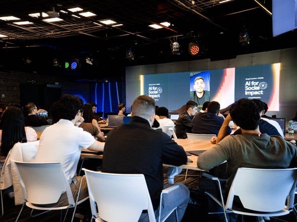 An audience is seated in a dimly lit room, watching a presentation on a large screen titled 'AI for Social Impact' featuring a remote speaker.