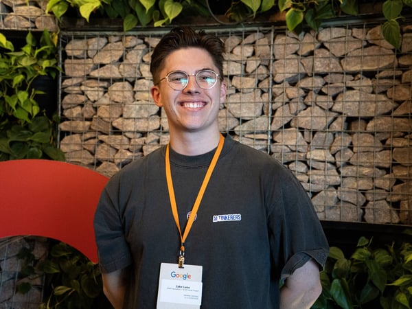 A portrait of a smiling young man wearing glasses and a Google event badge, standing in front of a stone gabion wall with green plants.