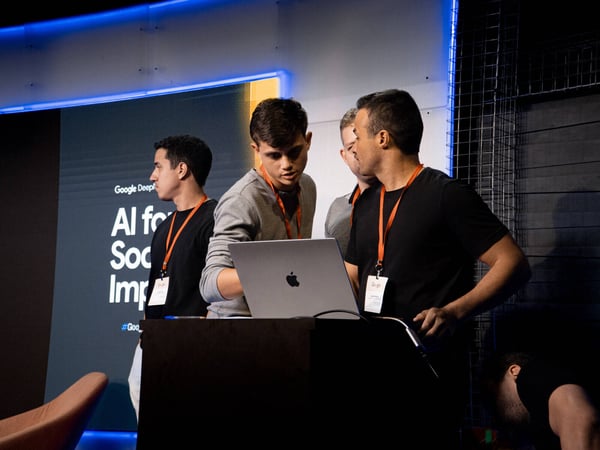 A group of men are gathered around a laptop on a podium at a Google DeepMind event titled 'AI for Social Impact'.