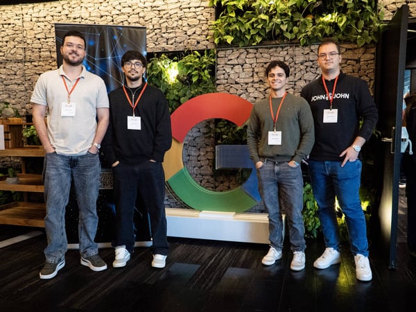Four young men are posing for a photograph in front of a large, multi-colored Google 'G' logo in an indoor setting with a stone wall and greenery.
