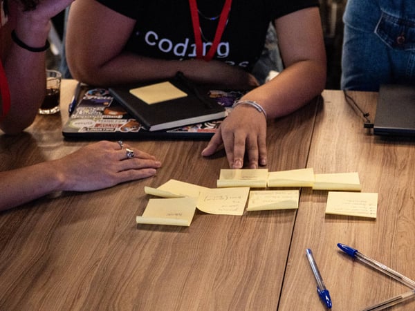 A close-up photograph of people collaborating around a wooden table, featuring yellow sticky notes with handwritten notes, pens, and a laptop decorated with stickers.