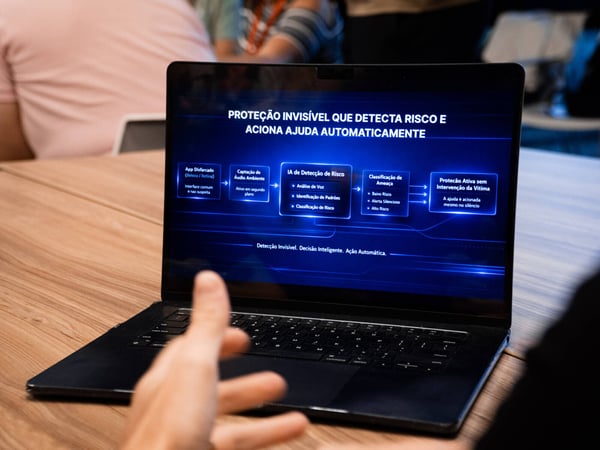 A photo of a laptop screen displaying a presentation in Portuguese about an AI-based security system, with people in the background.