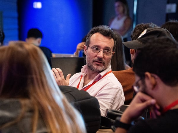 A man with glasses and a beard is engaged in a conversation during an indoor event, surrounded by other attendees.
