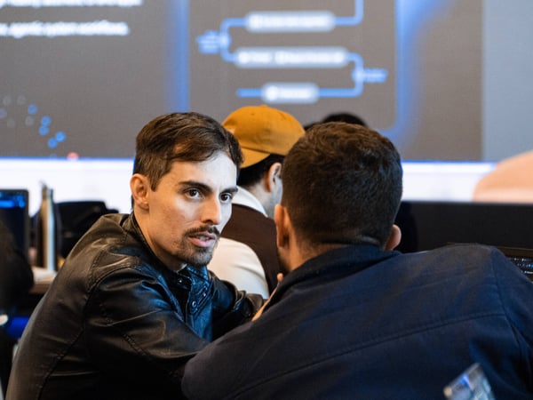 A man in a black leather jacket is engaged in a conversation with another person in an indoor setting, with a presentation screen displaying a flowchart in the background.