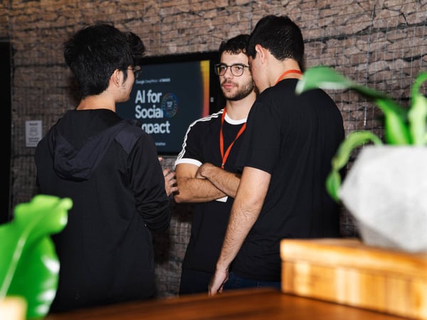 Three young men are engaged in a conversation at an event, standing in front of a screen displaying 'AI for Social Impact'.