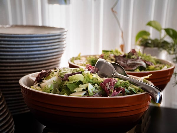 A close-up photograph of two large orange bowls filled with fresh salad, set next to a tall stack of plates in a catering or buffet setting.