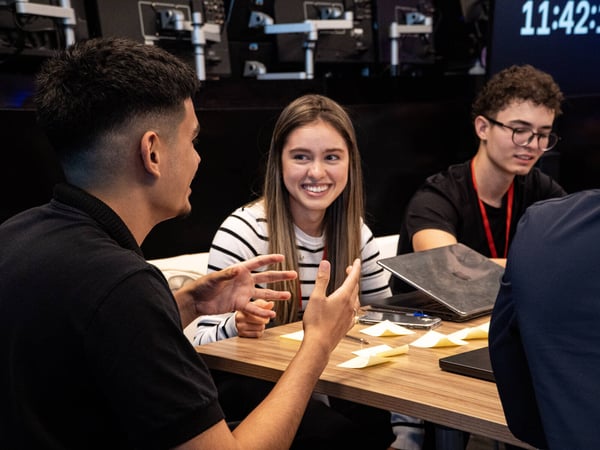 A group of young adults are engaged in a collaborative discussion around a wooden table with laptops and sticky notes.