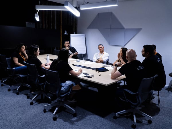 A group of eight people are gathered around a large conference table in a modern, dimly lit office for a meeting or discussion.