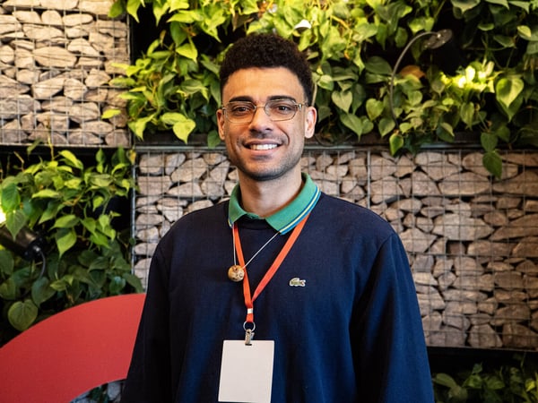 A portrait of a smiling man wearing glasses and a navy blue sweatshirt with a red lanyard, standing in front of a wall decorated with rocks and green plants.