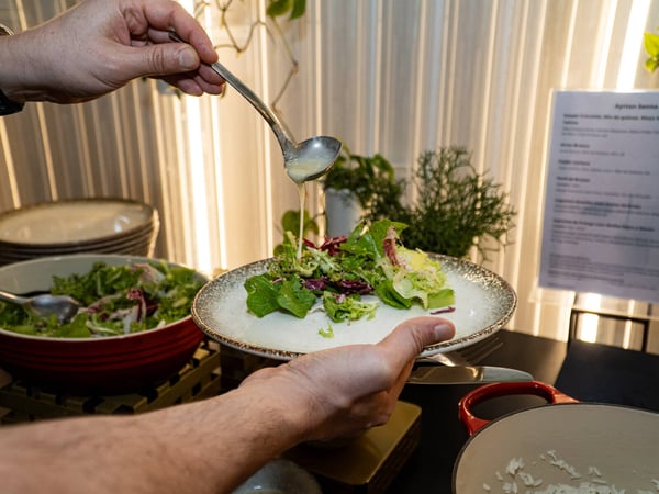 A close-up photo of a person pouring dressing from a small ladle onto a fresh salad on a plate, likely at a buffet or catered event.