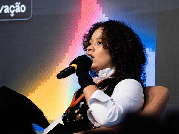 A woman with curly hair is speaking into a microphone while sitting on a stage during an event.