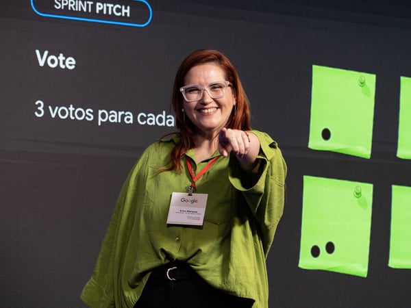 A woman with red hair and glasses smiles and points at the camera while standing in front of a presentation screen at a Google event.