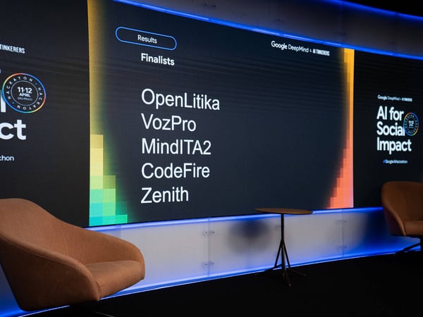 A photograph of a modern event stage featuring a large digital screen displaying a list of finalists for an 'AI for Social Impact' hackathon.