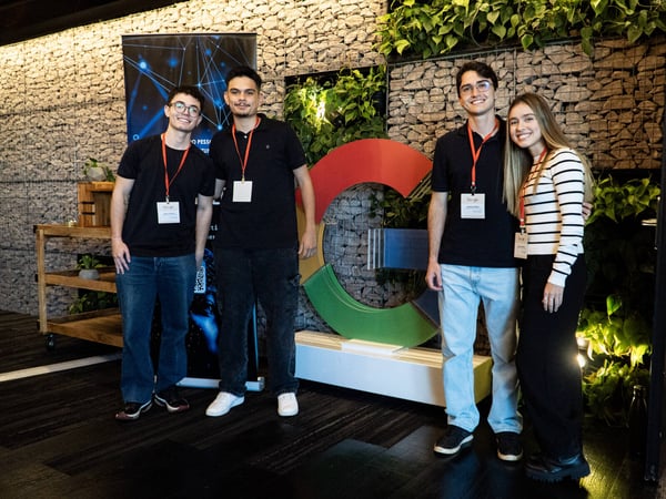 Four young people are posing for a group photo in an indoor setting featuring a stone wall, plants, and a large Google logo.
