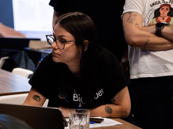 A woman with glasses and tattoos sits at a desk with a laptop, looking intently to the side, while others stand nearby in an office setting.
