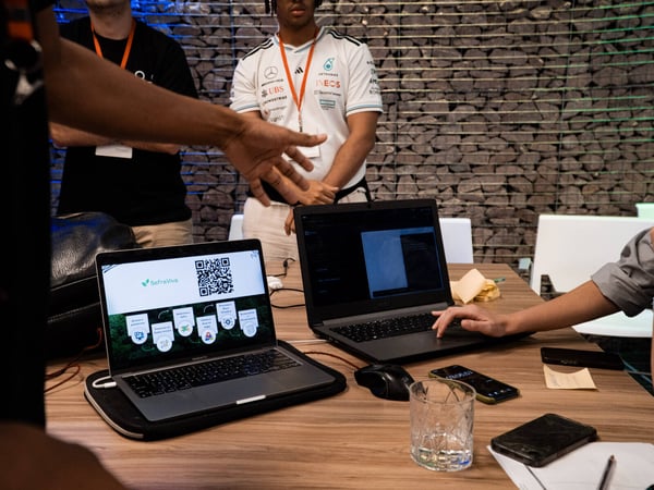 A group of people are gathered around a wooden table with laptops, one of which displays a website called 'SafraViva' with a QR code.