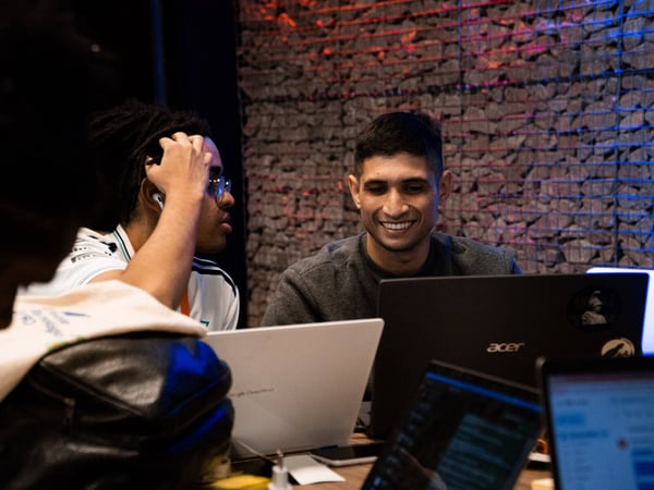 A group of young men are working on their laptops in a modern indoor setting with a stone-textured wall in the background.