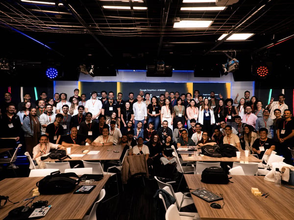 A large group of people posing for a group photo in a studio or event space with stage lighting and wooden tables in the foreground.