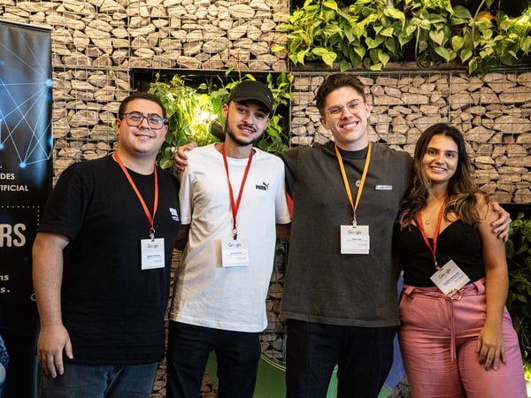 A group of four young adults, three men and one woman, posing for a photo in front of a stone wall and green plants, all wearing Google event lanyards.