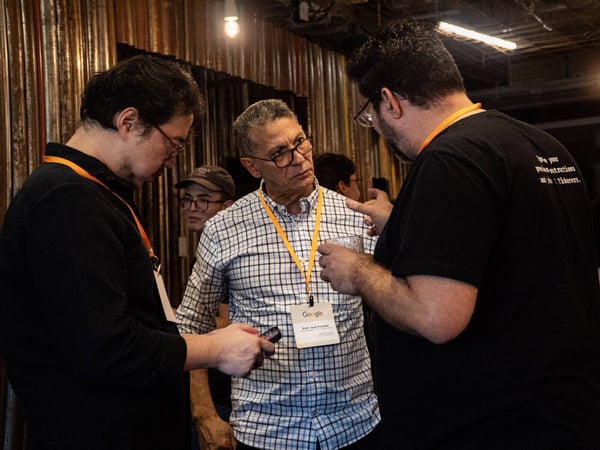 A group of men are engaged in a conversation at an indoor event, with one man wearing a Google lanyard and another wearing a t-shirt that mentions 'Tinkerers'.
