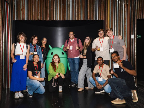A group of eleven people are posing for a photo around a large green Android mascot statue in an indoor setting with a textured wall.