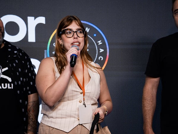 A woman with glasses and an orange lanyard speaks into a microphone during an indoor event, standing between two other people.