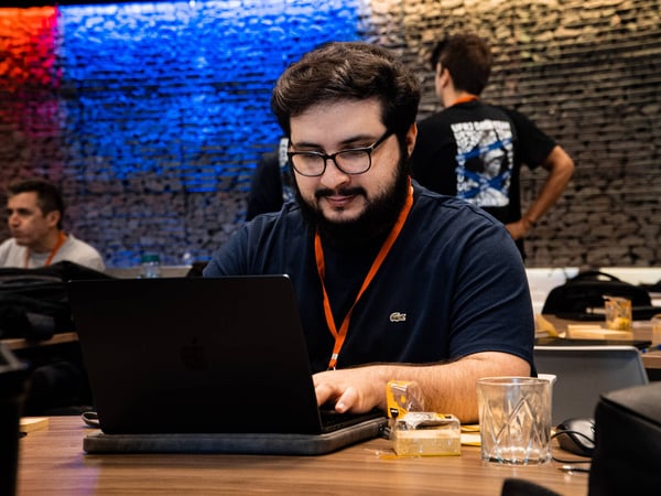 A man with a beard and glasses is focused on working on a laptop at a wooden table in an indoor setting with a textured stone wall background.