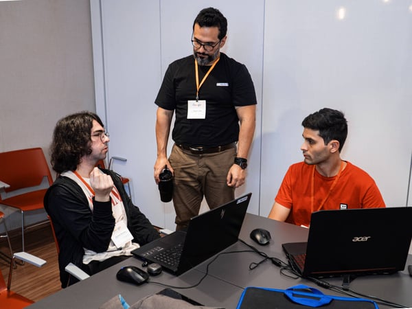 Three men are gathered around a table with laptops in an indoor setting, appearing to be engaged in a technical discussion or collaboration.