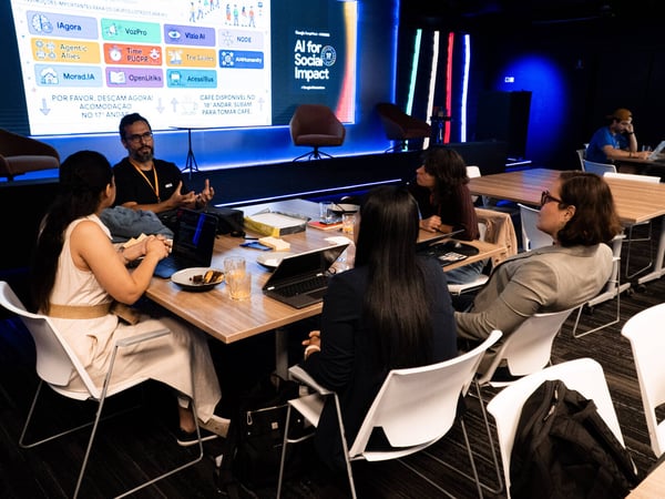 A group of people sitting around a wooden table in a modern office or event space, engaged in a discussion with a large digital screen in the background displaying event information.