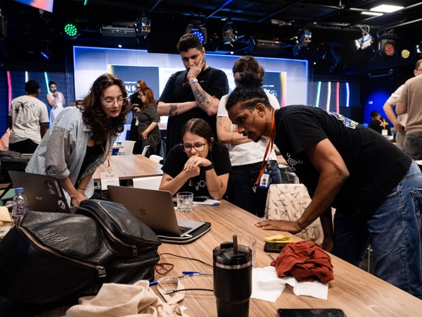 A group of people are gathered around a laptop on a wooden table, deeply engaged in a collaborative discussion during a tech event.
