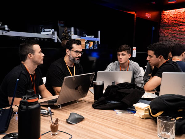 A group of men are seated around a wooden table with laptops, engaged in a focused discussion in a modern indoor workspace.