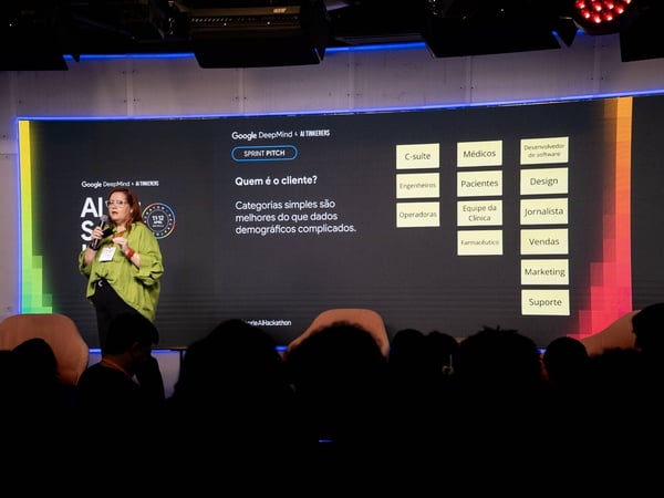 A woman in a lime green shirt is delivering a presentation on a stage in front of a large screen displaying a slide in Portuguese.