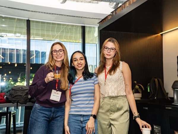 Three young women posing together indoors, likely at a professional event, wearing lanyards and holding drinks.