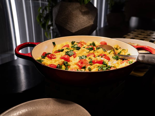 A close-up photograph of a red enameled pan filled with scrambled eggs, cherry tomatoes, and green leafy herbs or vegetables.