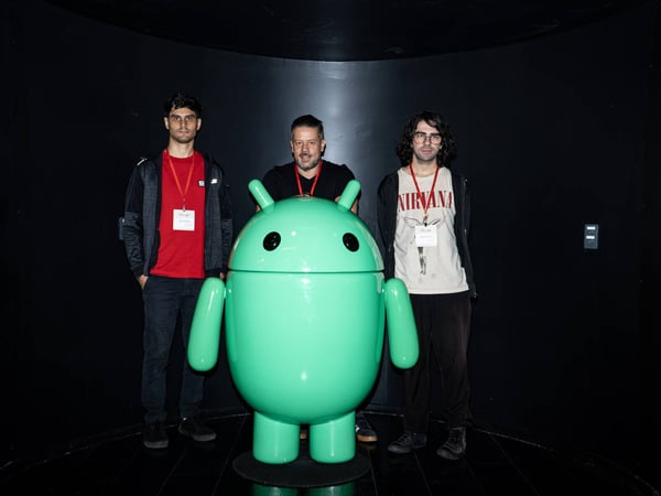 Three men are posing for a photo behind a large, green Android mascot statue in a dark, curved room.