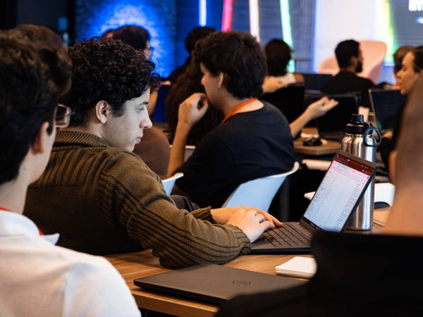 A young man with curly hair is focused on his laptop during a workshop or class, with several other participants visible in the background.