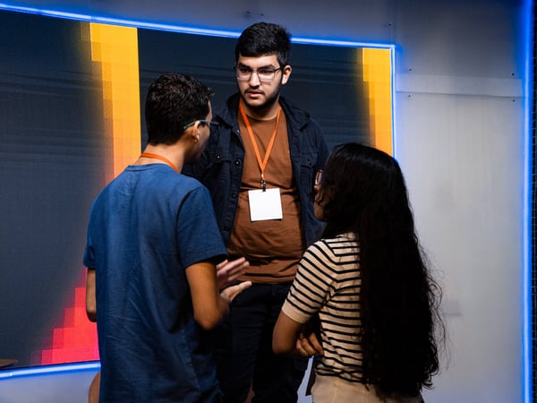 Three young people are engaged in a conversation at an indoor event, with one man facing forward and the others seen from the side or back.