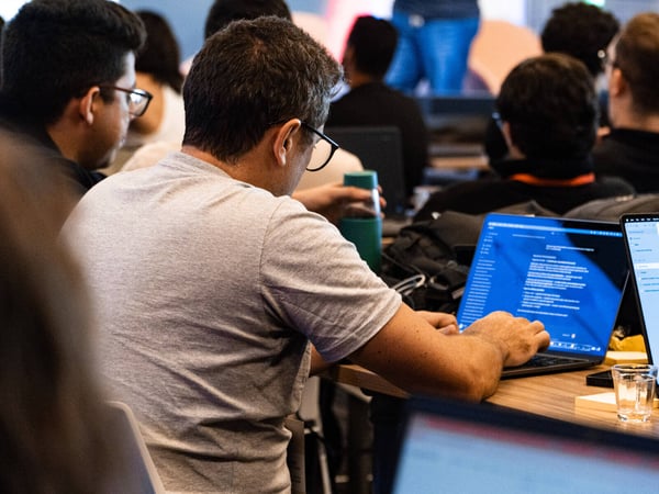 A group of people are seated at tables in an indoor setting, focusing on their laptops during a workshop or training session.