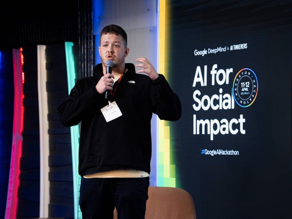 A man is speaking into a microphone on a stage in front of a screen displaying 'AI for Social Impact'.
