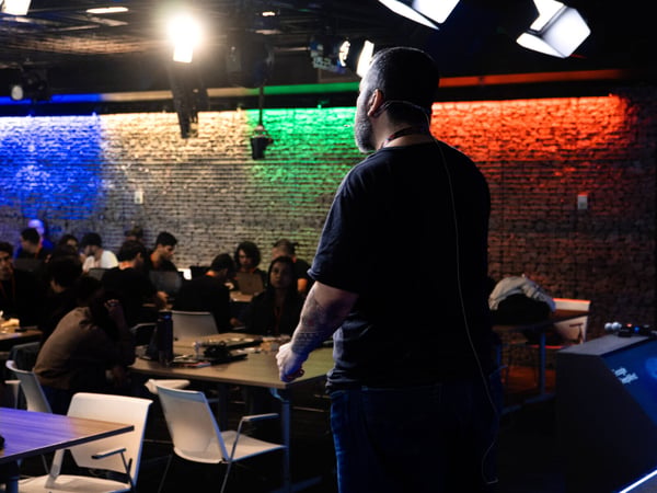 A speaker stands with his back to the camera, addressing an audience seated at tables in a room featuring a brick wall illuminated with blue, yellow, green, and red lights.