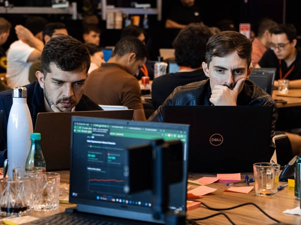 Two men are deeply focused on their laptops in a crowded, indoor workspace, likely during a hackathon or a collaborative project.