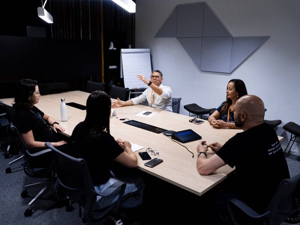 A group of five people are gathered around a large conference table in a modern office setting, engaged in a discussion led by a man gesturing at the head of the table.