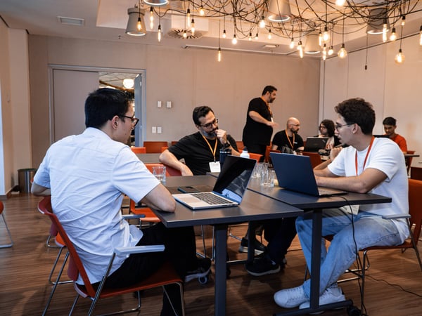 A group of people are engaged in a collaborative discussion or work session in a modern office space characterized by unique hanging light bulbs.