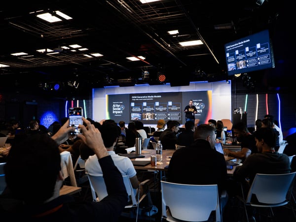A photograph of a presentation taking place in a large, dimly lit event space where an audience is watching a speaker on stage discussing generative media models.