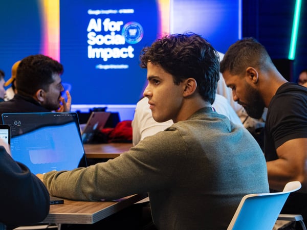 A young man with dark curly hair sits at a table with a laptop, focused on a presentation or discussion in a tech-oriented environment.