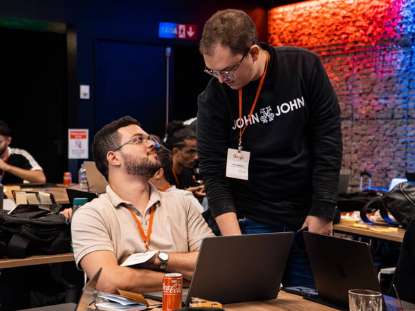 Two men are engaged in a discussion at a tech event or workspace, with one man standing and leaning over to talk to another who is seated with a laptop.